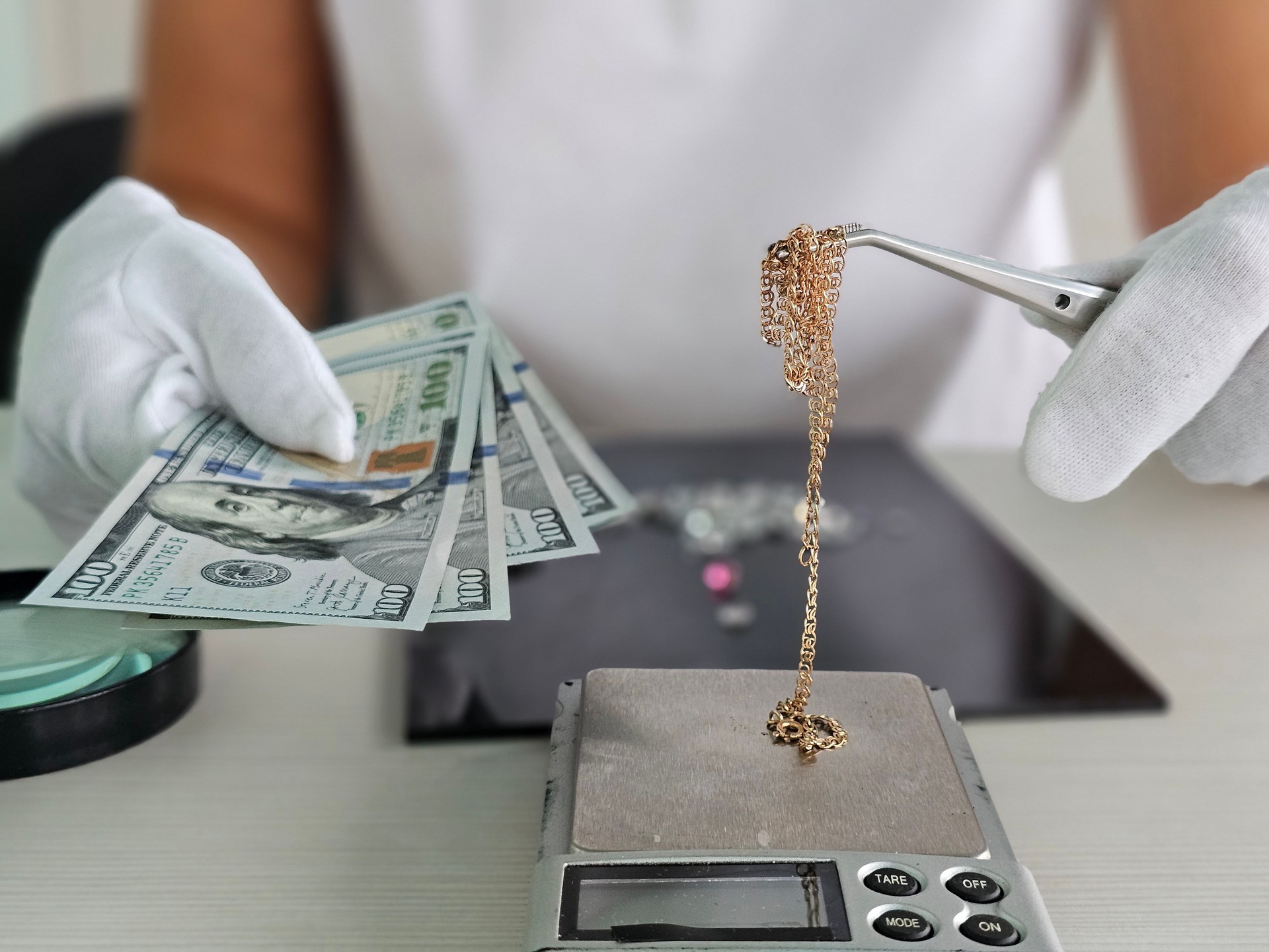 A jeweler weighs a gold chain on a precision scale while holding several banknotes in his other hand in a jewelry store concept closeup